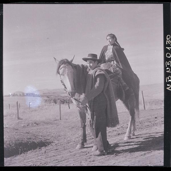 [Mujer mapuche a caballo, su marido tira las riendas por un camino rural]