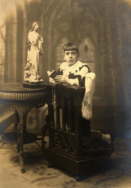 [Retrato de un niño en el día de su Primera Comunión, en sus manos sostiene un misal y rosario, se encuentra en un reclinatorio, junto a una mesita con la imagen de la Virgen]
