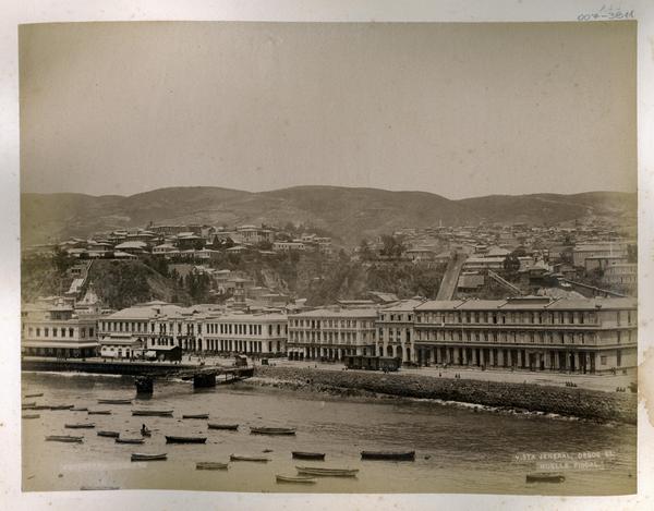 [Vista general de Valparaíso desde el muelle fiscal, en el mar se divisan botes y en el fondo lo cerros]